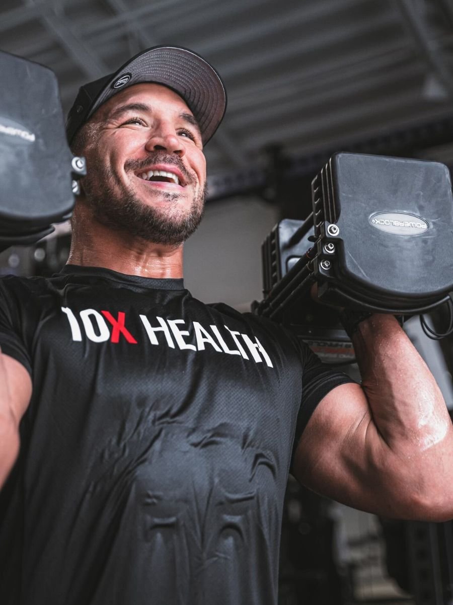 a smiling man lifting two dumbbells while wearing a black 10X Health t-shirt a smiling man lifting two dumbbells while wearing a black 10X Health t-shirt